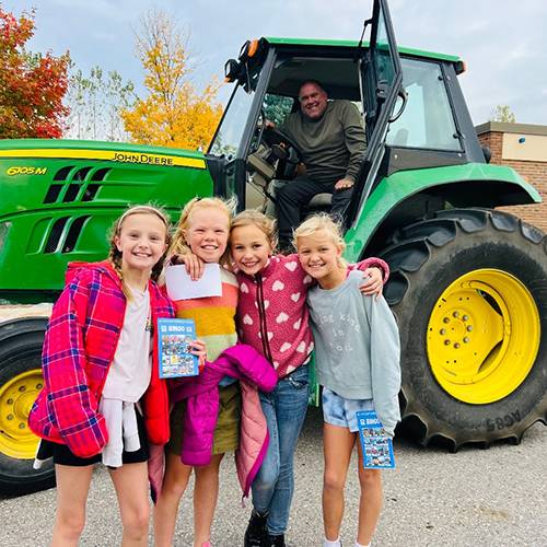 Four girls stand in front of a tractor while a farmer smiles from the tractor's cabin.